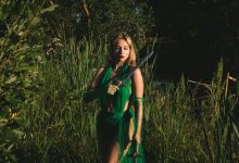 Mulher com vestido verde segura uma espada em uma floresta de atmosfera mística.