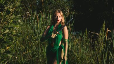 Mulher com vestido verde segura uma espada em uma floresta de atmosfera mística.