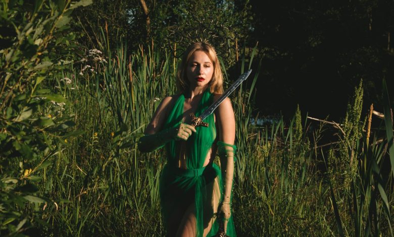 Mulher com vestido verde segura uma espada em uma floresta de atmosfera mística.