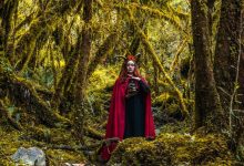 Mulher com capa vermelha segura um crânio em uma floresta densa e sombria.