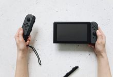 Mãos seguram um Nintendo Switch e controles Joy-Con sobre uma superfície branca.