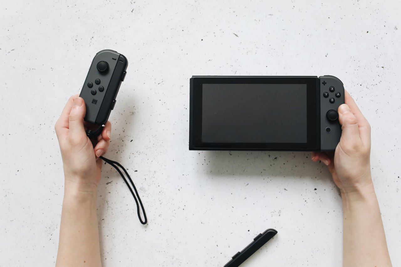 Mãos seguram um Nintendo Switch e controles Joy-Con sobre uma superfície branca.