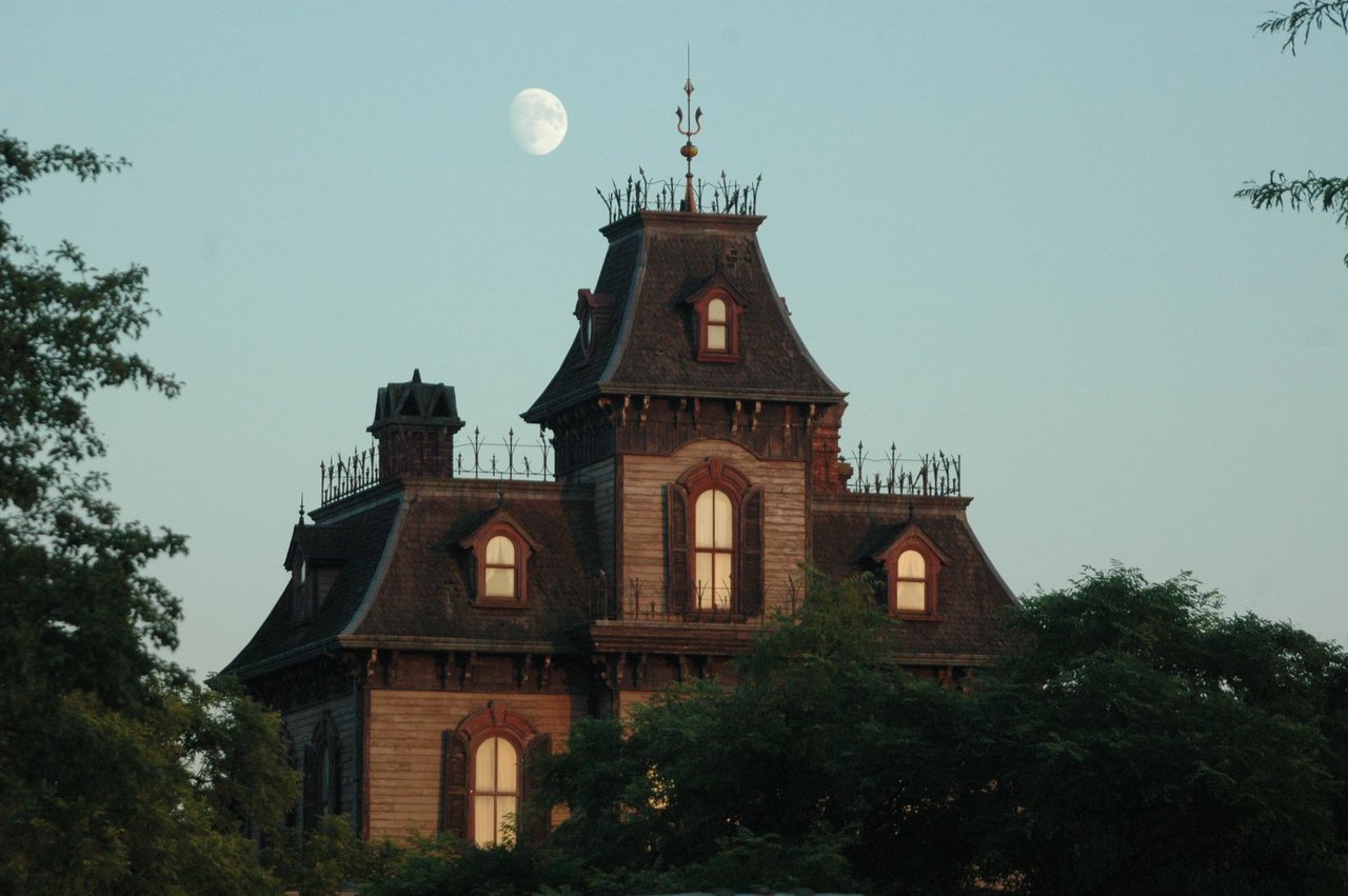 Mansão vitoriana sob o céu crepuscular, com a Lua visível ao fundo.