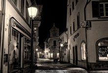 Rua de paralelepípedos entre prédios históricos, iluminada à noite.