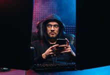 Homem usa um smartphone entre duas telas de computador em um ambiente escuro.