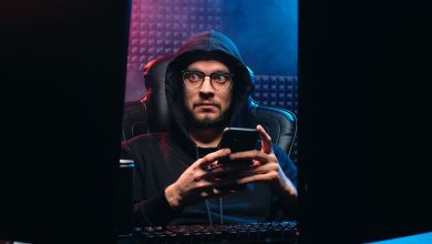 Homem usa um smartphone entre duas telas de computador em um ambiente escuro.