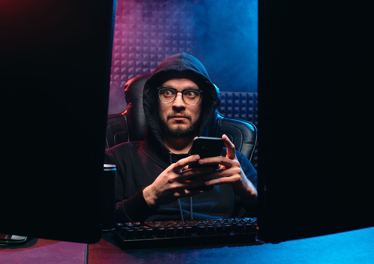 Homem usa um smartphone entre duas telas de computador em um ambiente escuro.