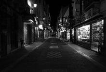 Rua urbana iluminada à noite, com vitrines comerciais, apresentada em preto e branco.