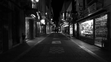 Rua urbana iluminada à noite, com vitrines comerciais, apresentada em preto e branco.