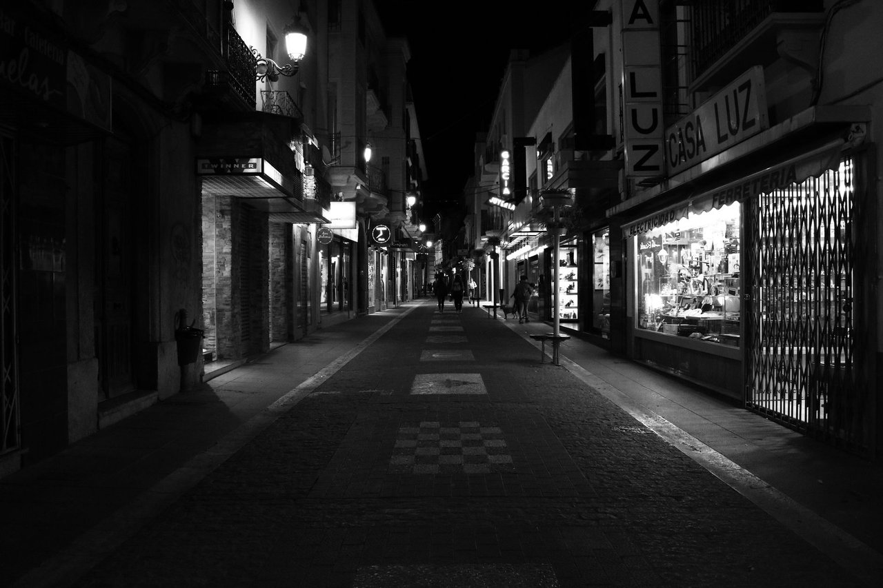 Rua urbana iluminada à noite, com vitrines comerciais, apresentada em preto e branco.
