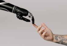 Mão humana tatuada se aproxima de uma mão robótica sobre fundo branco, simbolizando a relação entre humanos e inteligência artificial.