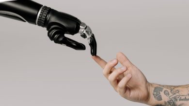 Mão humana tatuada se aproxima de uma mão robótica sobre fundo branco, simbolizando a relação entre humanos e inteligência artificial.