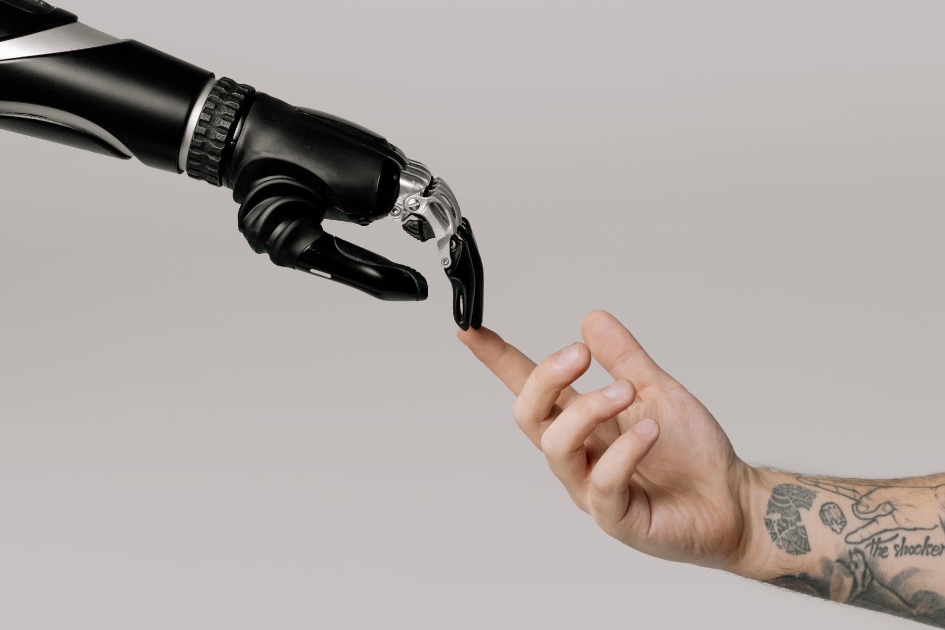 Mão humana tatuada se aproxima de uma mão robótica sobre fundo branco, simbolizando a relação entre humanos e inteligência artificial.