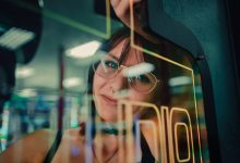 Mulher refletida no vidro de uma máquina de arcade, envolta por luzes neon.
