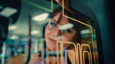 Mulher refletida no vidro de uma máquina de arcade, envolta por luzes neon.